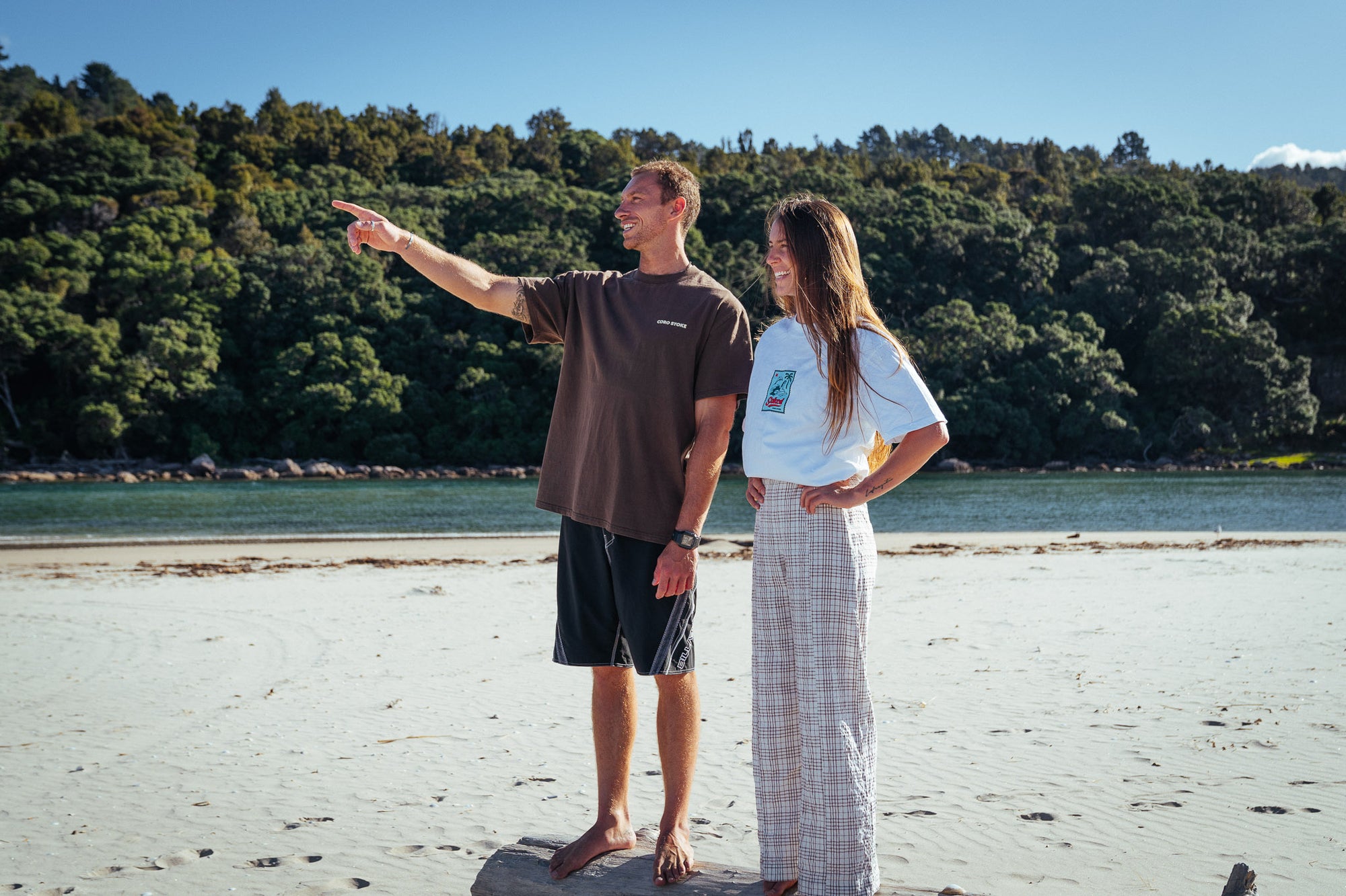 Two people standing on a beach with trees and water in the background. Both of them are wearing the new Coro Stoke Tee Designs.
