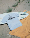 Light blue t-shirt with text on a white surfboard on a sandy beach.