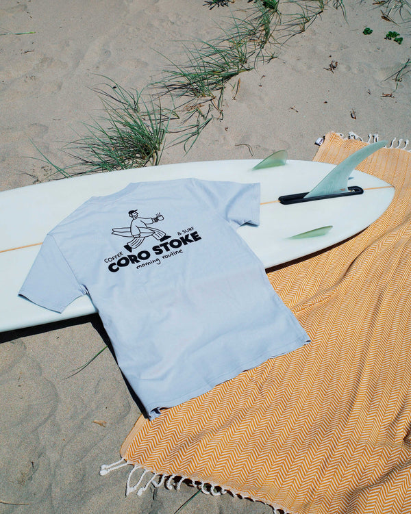 Light blue t-shirt with text on a white surfboard on a sandy beach.