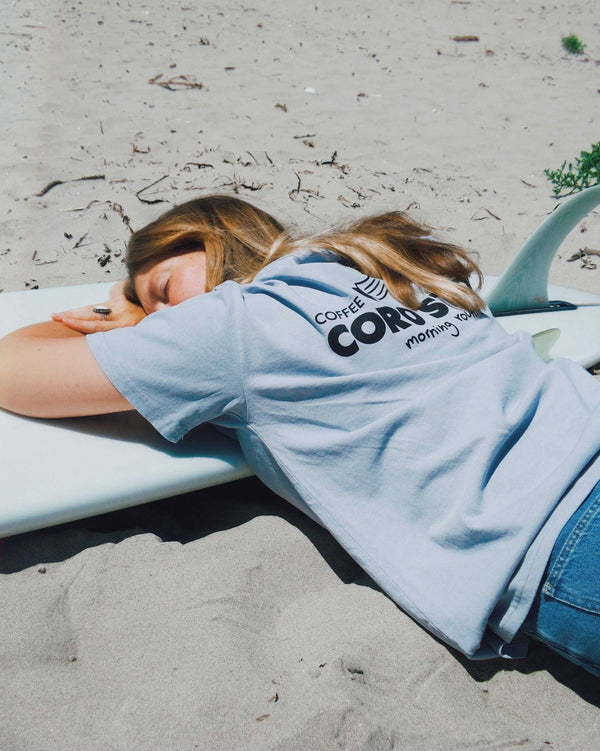 Person lying on a surfboard wearing a light blue t-shirt with text, on a sandy beach.