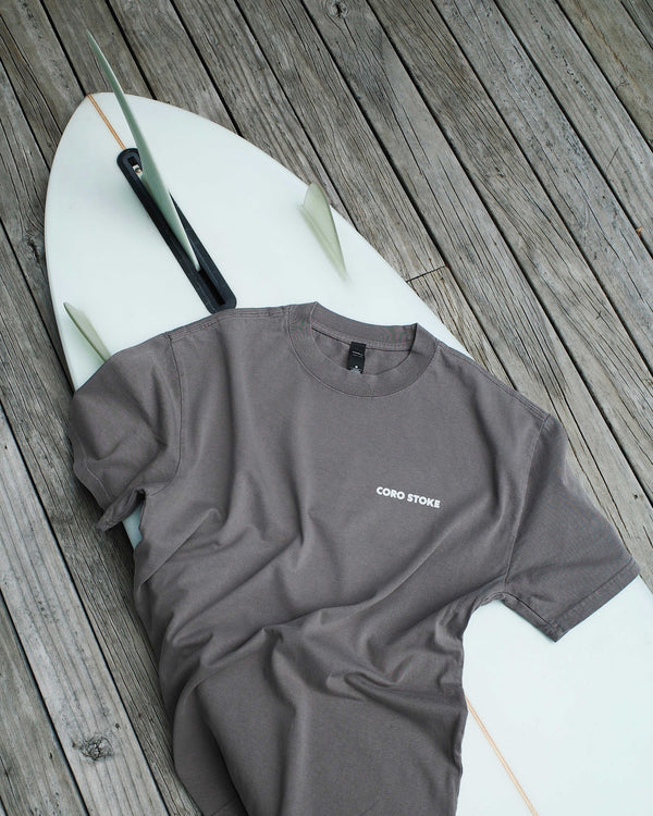 Gray t-shirt with 'Coro Stoke' logo on a white surfboard on a wooden deck