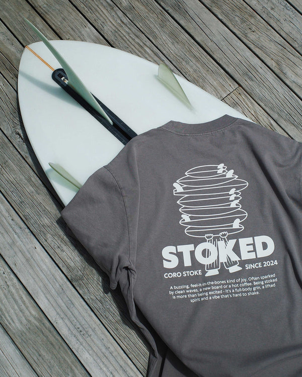 Gray t-shirt with 'STOKED' design on a wooden surface with a surfboard.