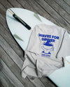 Gray tee with 'Waves for Dinner' design on a white surfboard with fins.