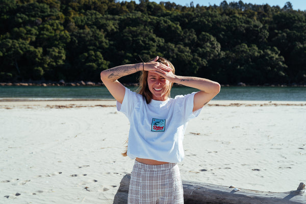 Person with the new Stamp Minus Tee from Coro Stoke is standing on a beach with trees and water in the background