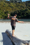 Person wearing the new Wave Therapy T-Shirt from Coro Stoke and walking on a log with arms outstretched on a sandy beach with greenery in the background