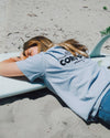 Person lying on a surfboard wearing a light blue t-shirt with text, on a sandy beach.