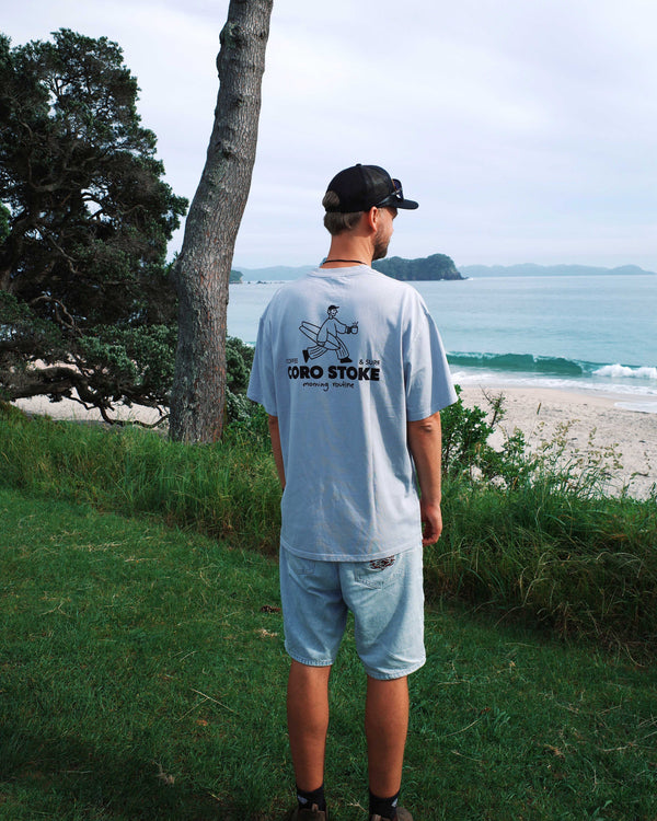 Person wearing a light blue t-shirt with a logo and text, standing on a grassy area with a beach and ocean view.