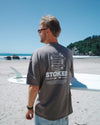 Man wearing a 'Stoked' t-shirt on a beach with a surfboard.