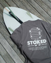 Gray t-shirt with 'STOKED' design on a wooden surface with a surfboard.