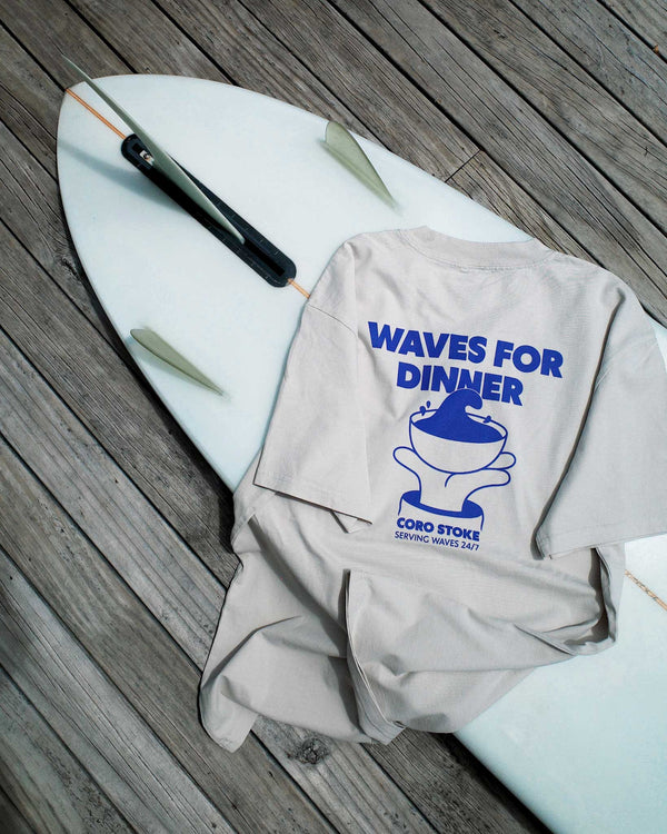 Gray tee with 'Waves for Dinner' design on a white surfboard with fins.