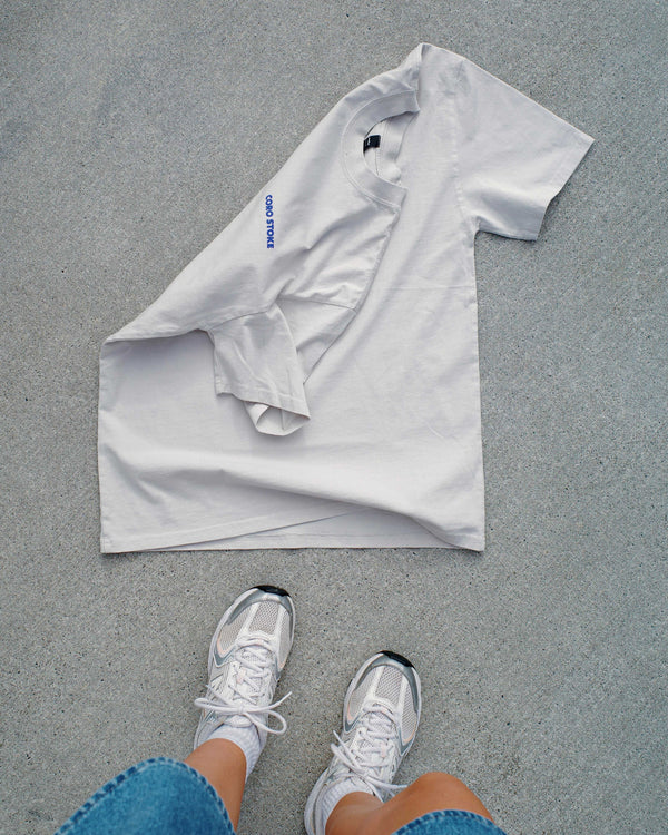 Grey shirt on a concrete surface with a person wearing white sneakers and blue jeans.
