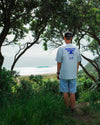 Person standing in a forested area looking towards a beach with 'Waves for Dinner' t-shirt.